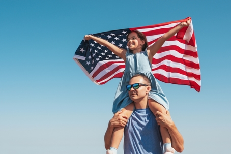 father carrying daughter on his shoulders who is holding the US flag